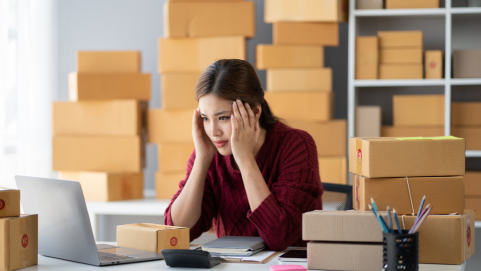 A frustrated person stares at their laptop, with boxes stacked on shelves behind them