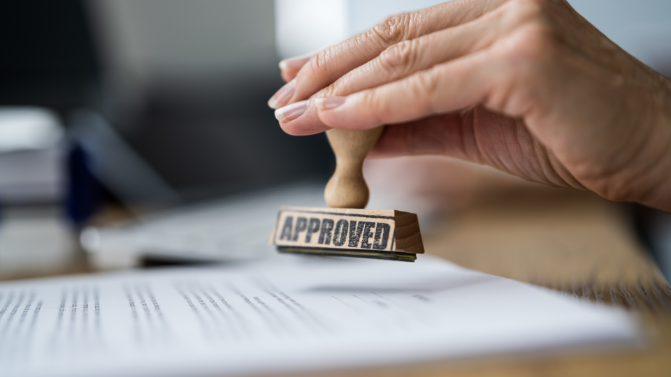 A rubber stamp with the word “approved” held above a piece of paper