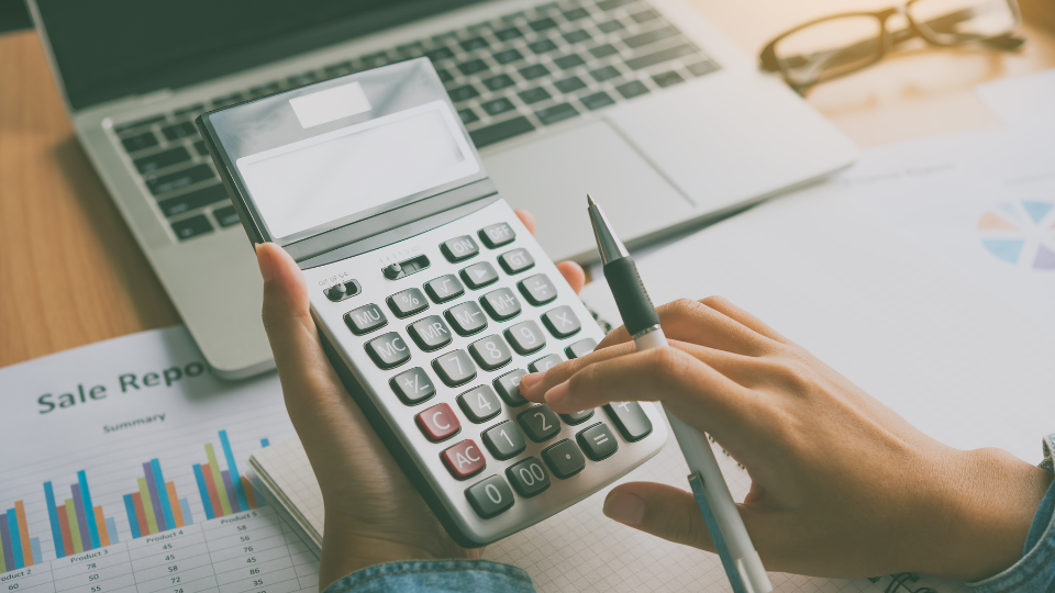 A person using a calculator on a desk in front of a laptop