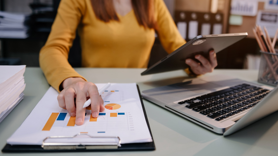 A person sitting at a desk with a laptop and clipboard monitoring market trends