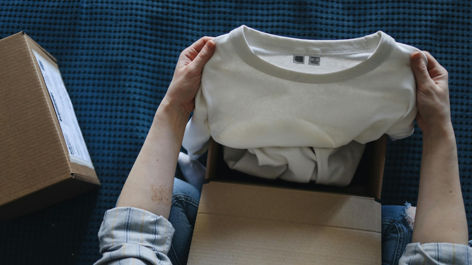 A person sitting on a bed opening a box with a t-shirt inside