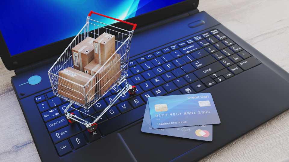 A mini shopping cart filled with boxes sits on top of a laptop next to two credit cards